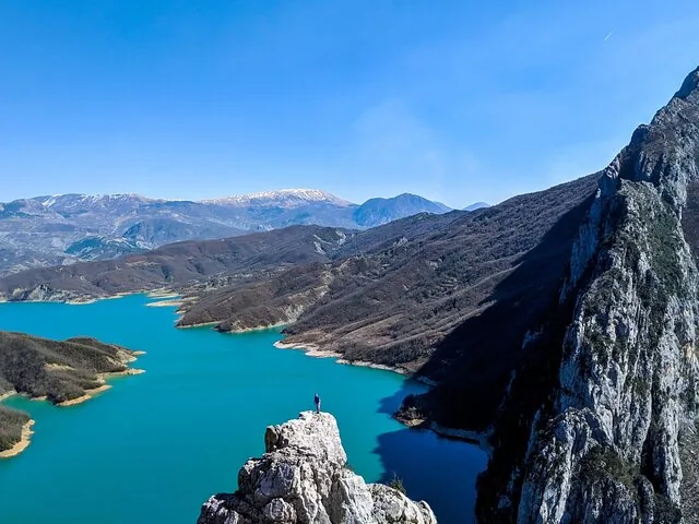 A person stands on a rocky peak overlooking Bovilla Lake and Gamti Mountain.