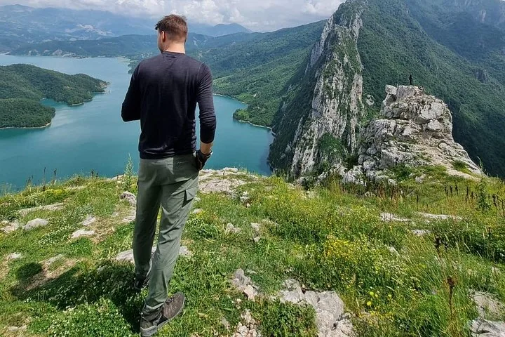 Ein Mann steht auf einer grasbewachsenen Klippe mit Blick auf den Bovilla-See und den Berg Gamti.