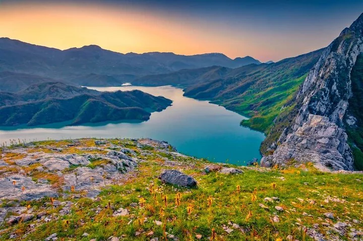 Sonnenuntergang am Bovilla-See mit schroffen Klippen und Bergen im Hintergrund.